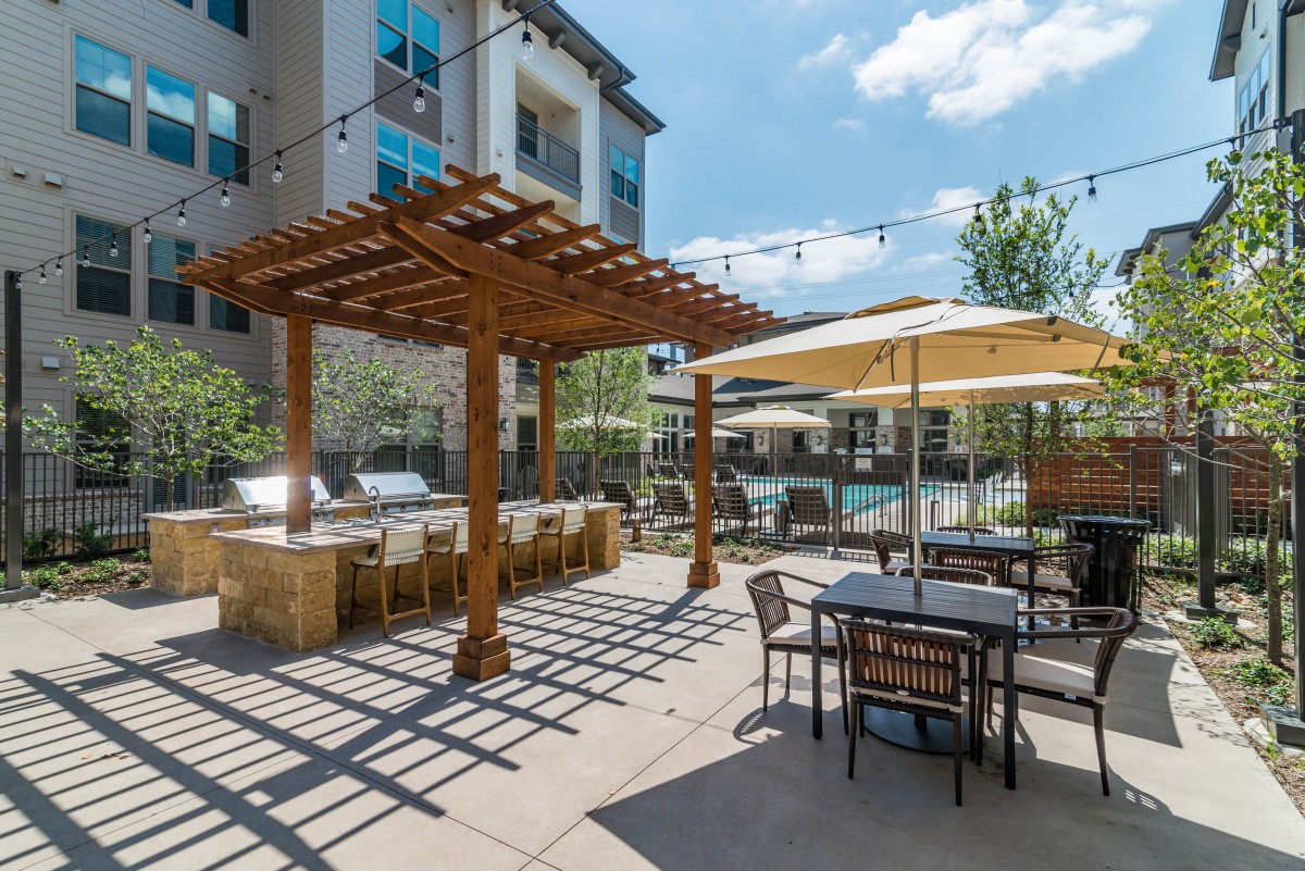An outdoor patio area with a pergola, tables, chairs, and umbrellas on a sunny day.