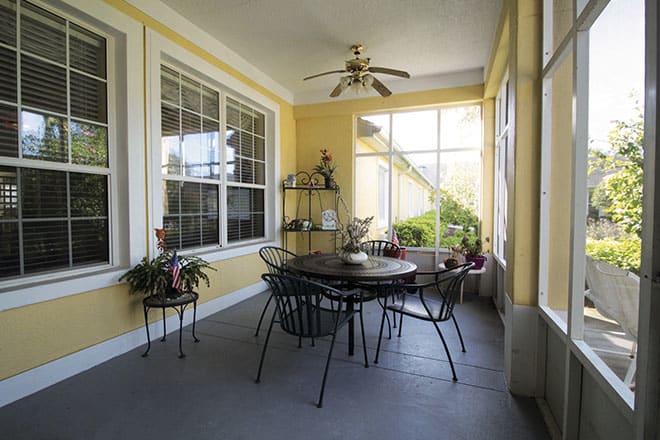 A cozy porch with a round table and chairs, surrounded by windows and adorned with plants.