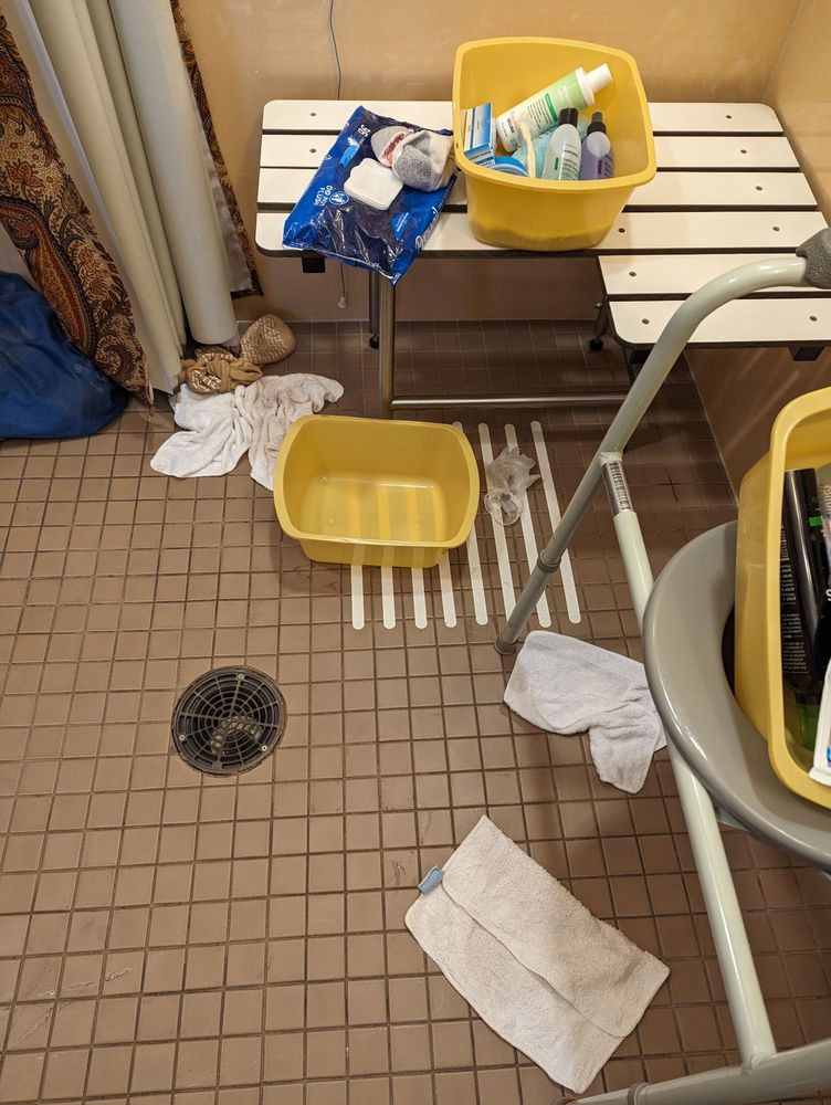 A bathroom with cleaning supplies and a yellow bucket on a tiled floor.