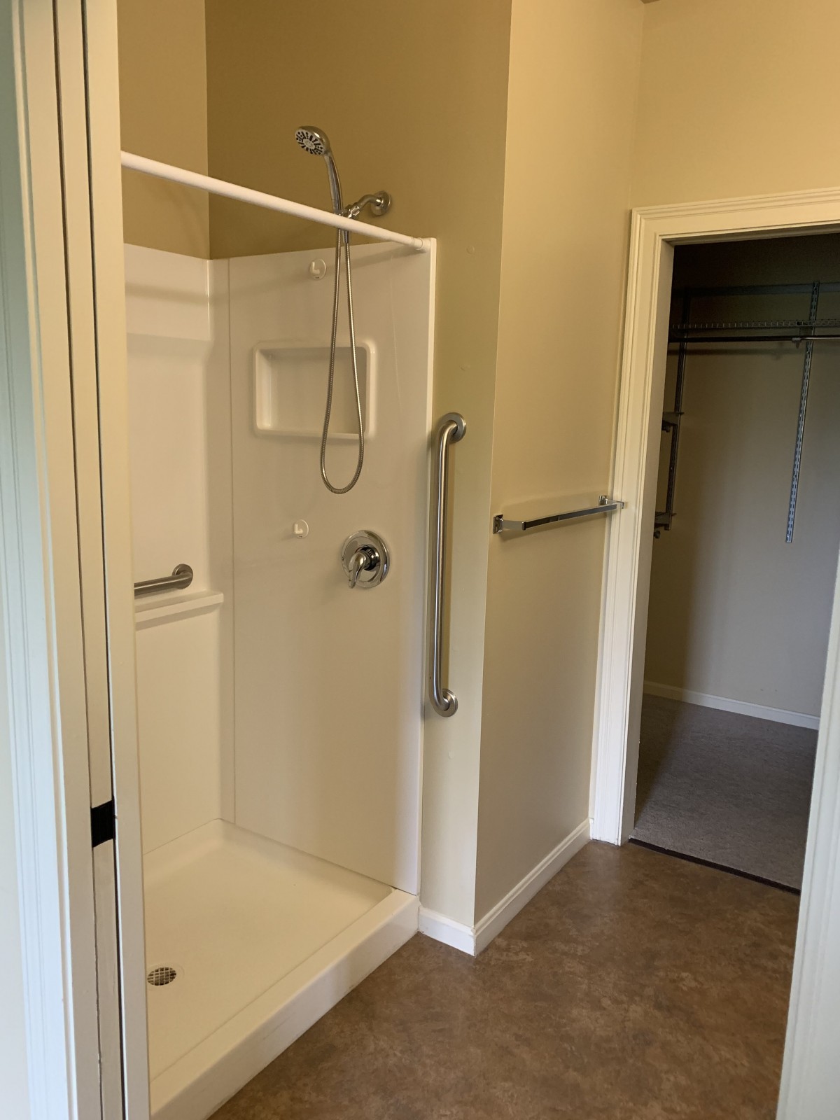 A modern walk-in shower with grab bars and an adjacent closet.