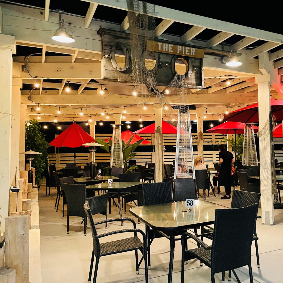 An outdoor dining area with tables and chairs under red umbrellas, illuminated by string lights at night.