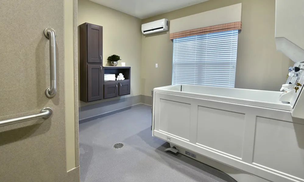 A modern bathroom interior featuring a walk-in bathtub, cabinetry, and window blinds under an air conditioning unit.