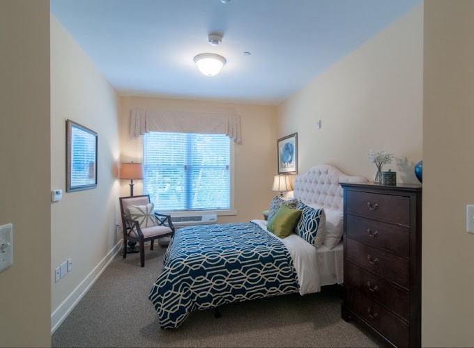 A cozy bedroom with a neatly made bed, adorned with decorative pillows, flanked by lamps. The room has an elegant chair and a window allowing natural light.