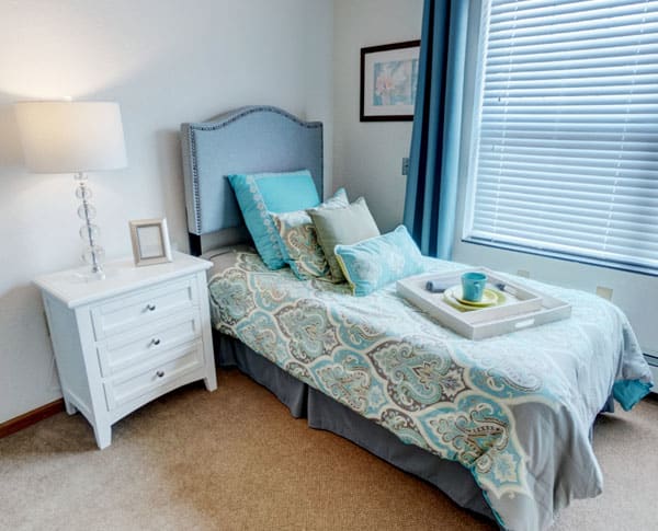 A cozy bedroom with a neatly made bed, featuring pastel-colored pillows and a decorative tray, alongside a white nightstand with a lamp.