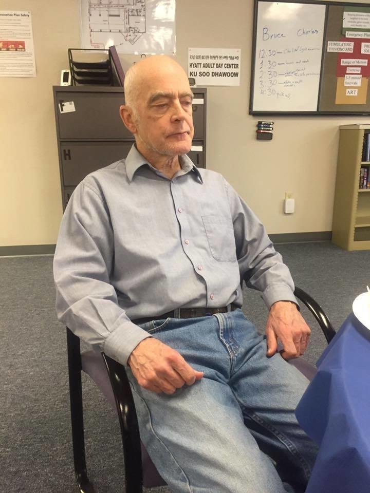 An elderly man sitting in an office environment, wearing a shirt and jeans.