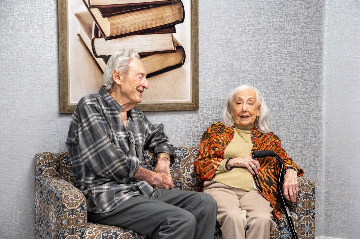 An elderly couple sitting together, engaged in conversation beside a painting of books.