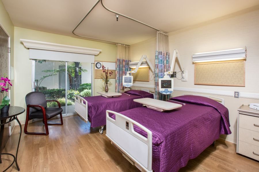 A modern hospital room featuring twin beds with medical equipment and a view of greenery outside.