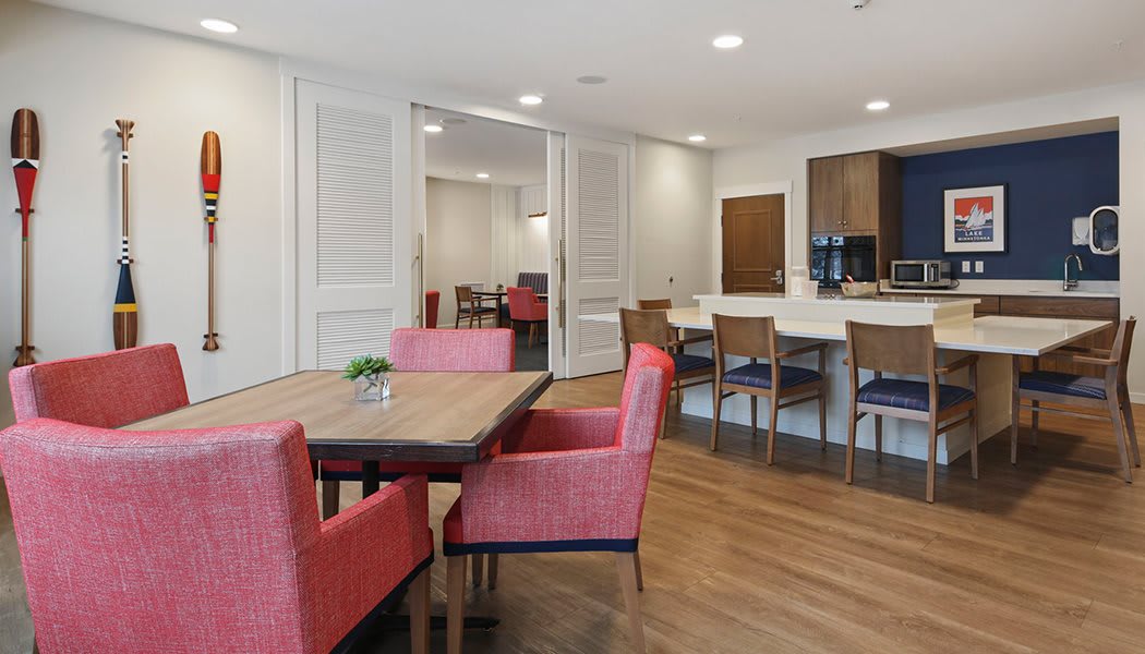 A modern dining area with a wooden table, surrounded by red chairs, adjacent to a kitchen counter with bar stools and wood flooring.