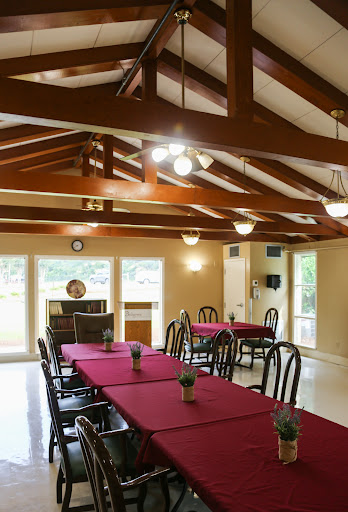 A cozy dining area featuring tables dressed with red tablecloths, wooden beams overhead, and ample natural light.