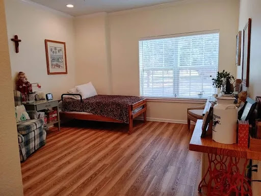 A cozy and simple room with a bed, a small table, and chairs, featuring natural light from a window.