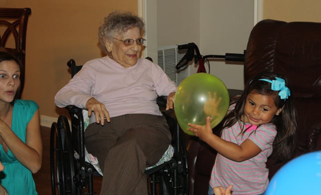 An elderly woman sitting in a wheelchair smiles while watching a child play with a balloon.
