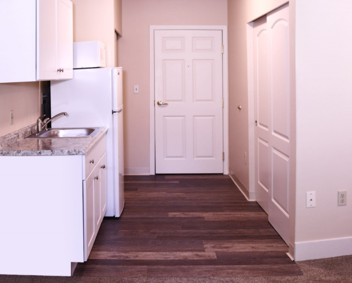 A compact apartment kitchen with white cabinets and a refrigerator, featuring wooden flooring.
