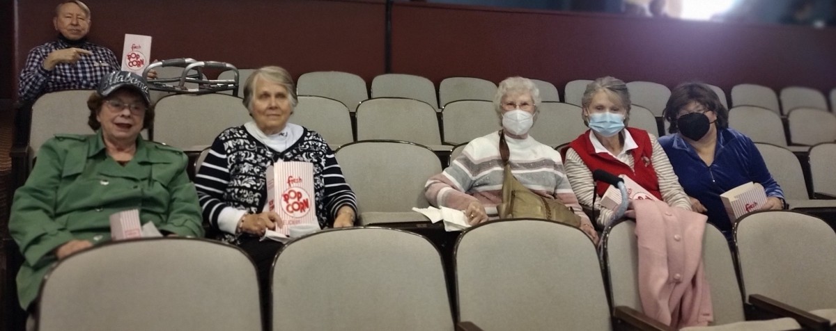 Three elderly women sitting in a theater, enjoying popcorn with one wearing a mask.