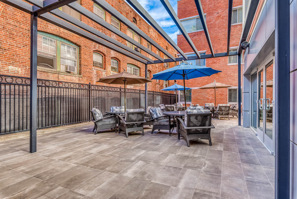 A modern outdoor patio area with tables, chairs, and umbrellas, set against a brick building backdrop.