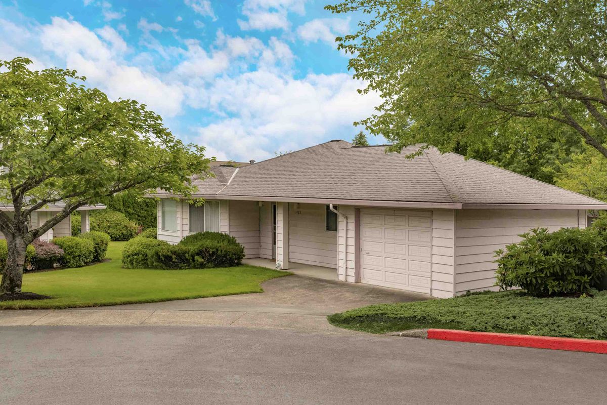 A suburban house with a front yard and driveway, surrounded by trees under a partly cloudy sky.