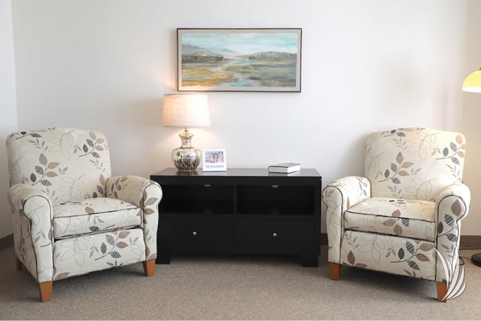 A cozy living room setup featuring two floral armchairs, a dark side table with a decorative lamp, and a landscape painting on the wall.