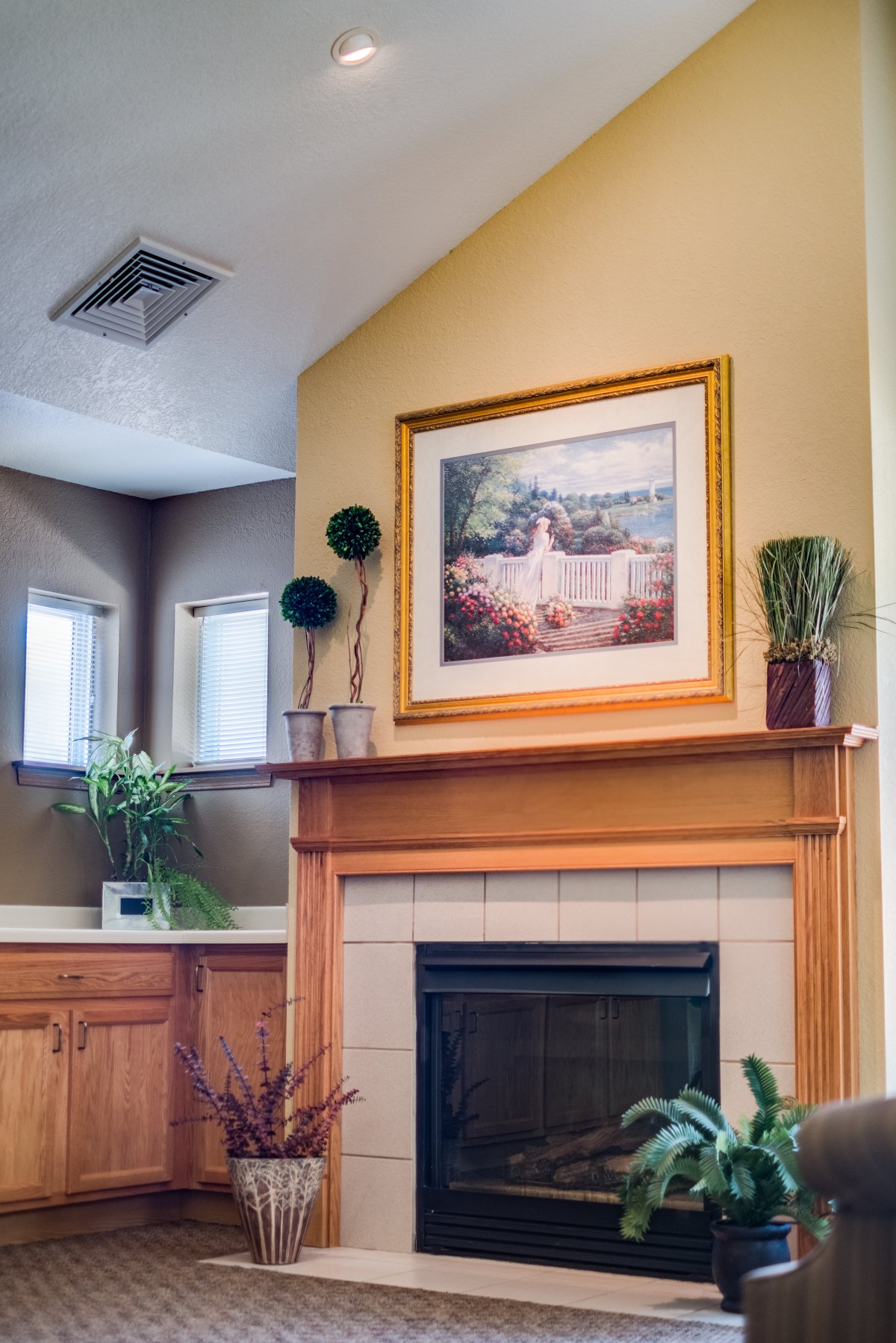 A cozy living room with a fireplace, adorned with plants and a framed painting above the mantel.
