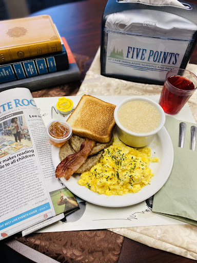 A hearty breakfast spread featuring scrambled eggs, toast, bacon, grits, and a side of newspaper reading.