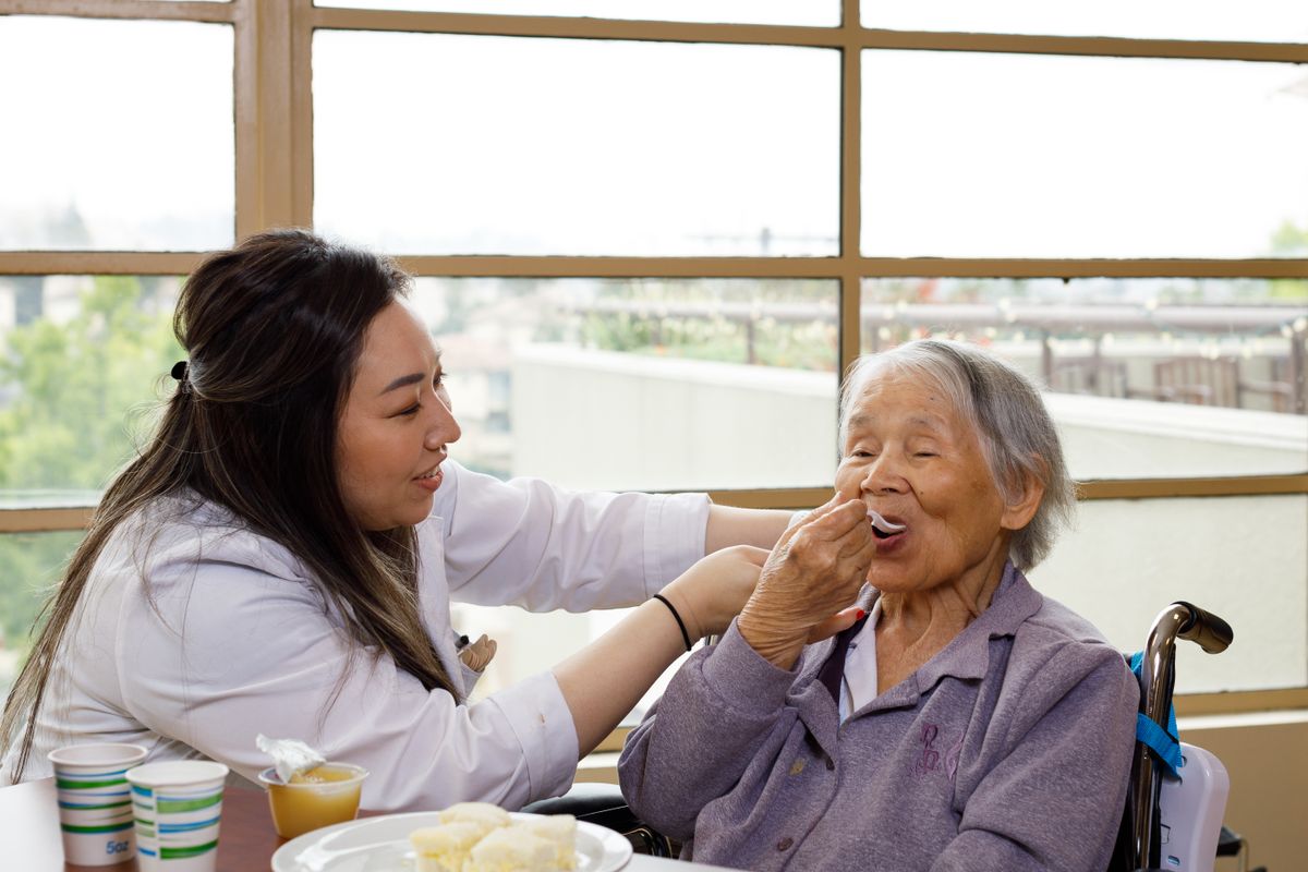 A caregiver assists an elderly woman by feeding her in a comfortable setting.