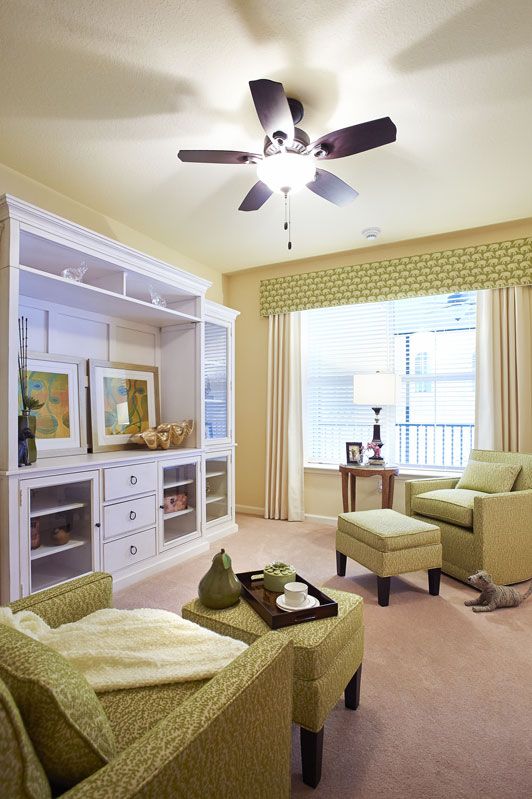 A cozy living room with light green armchairs and a well-organized bookshelf, illuminated by natural light from a large window.
