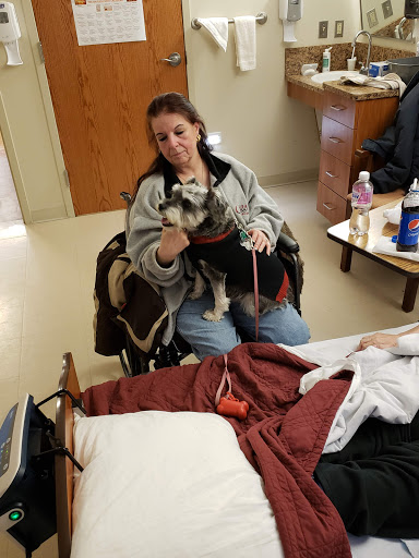 A woman in a wheelchair holding a small dog during a visit in a hospital room.