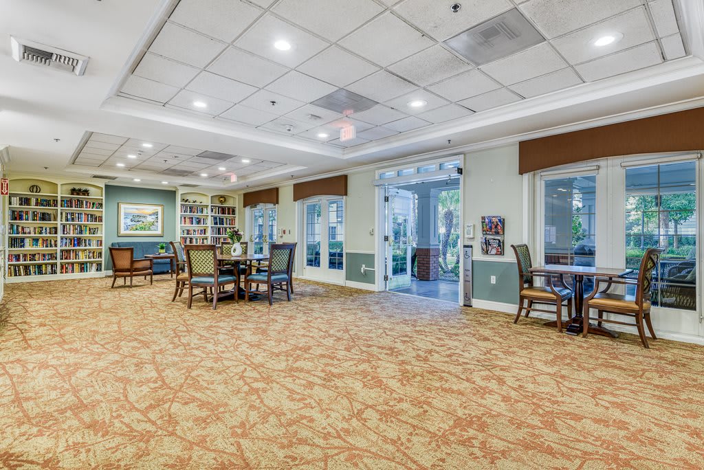 A spacious, well-lit lounge area with tables and chairs, featuring large windows and a carpeted floor.