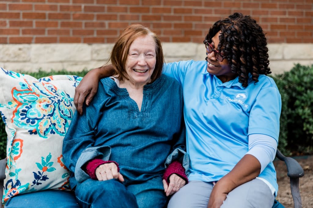 An elderly woman and her caregiver share a moment of laughter and companionship outdoors.