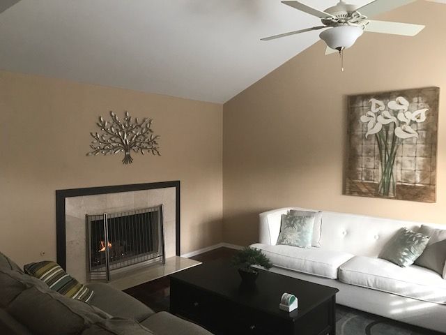 A cozy living room with a modern aesthetic, featuring a sofa, fireplace, ceiling fan, and decorative wall art.