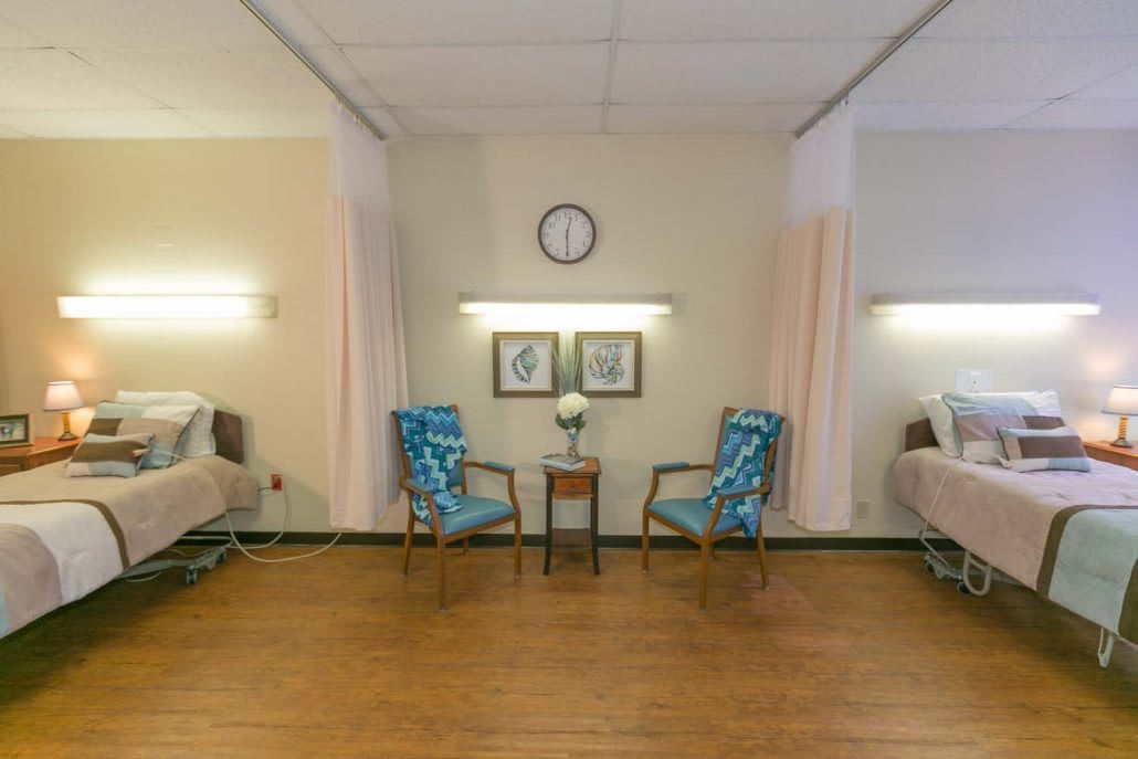 A hospital room with two beds separated by curtains, featuring chairs and a small table in between.