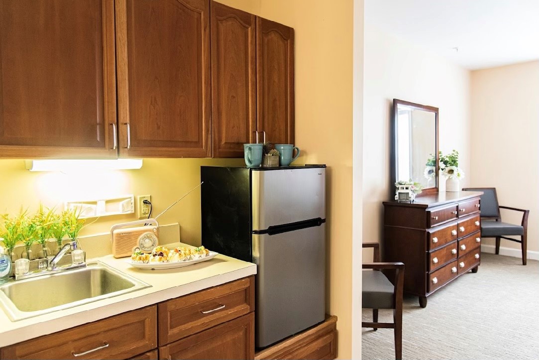 A cozy kitchenette with brown cabinets and a mini fridge next to a sink, leading into a room with a dresser.