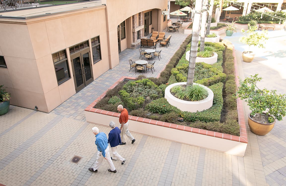A courtyard with people walking along a paved path, surrounded by garden landscape and outdoor seating.
