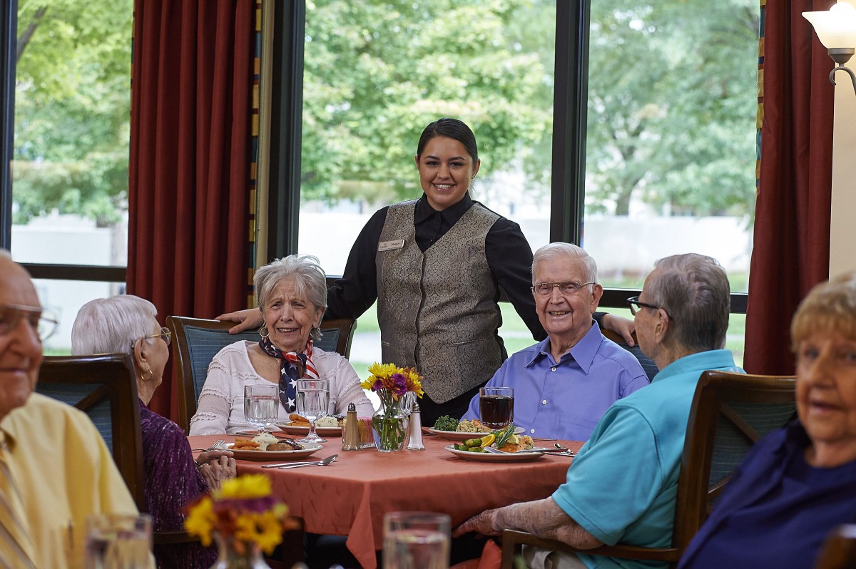 A group of elderly people enjoying a meal together in a dining area with a server standing nearby.