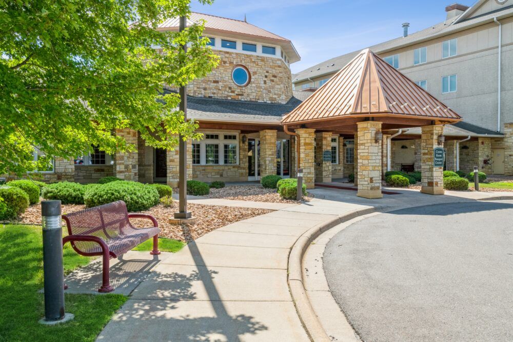 A stone building with a covered entrance, surrounded by landscaped greenery and a pathway with a bench.