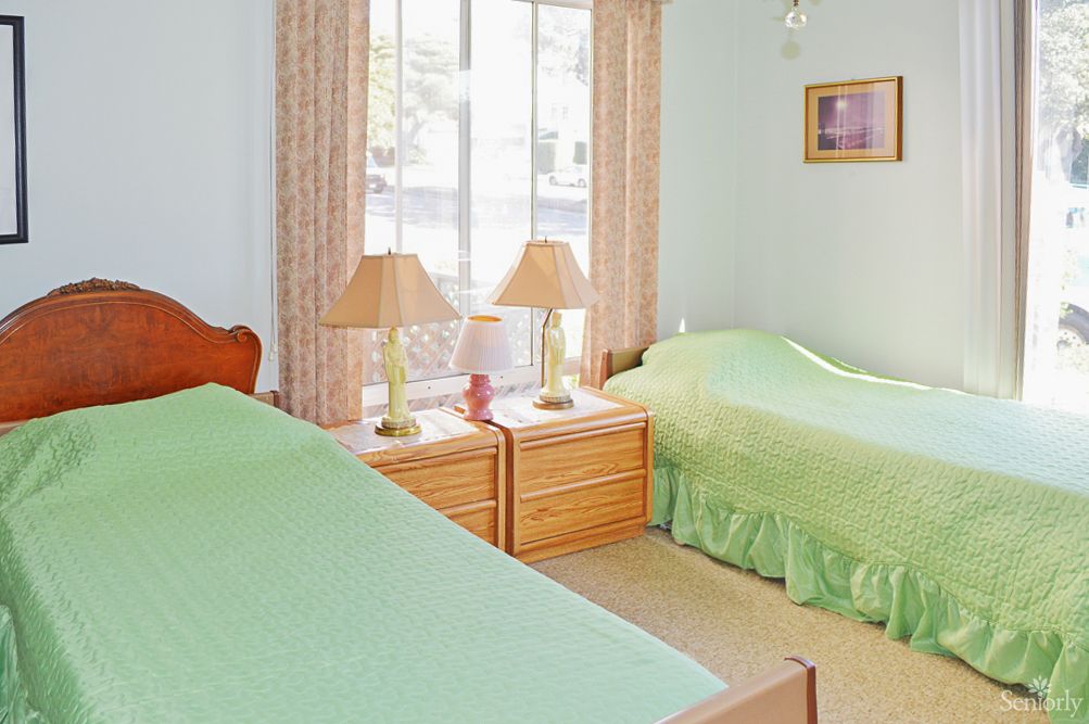 A cozy bedroom featuring two twin beds with green bedspreads, complemented by matching lamps on a wooden nightstand next to a large window.
