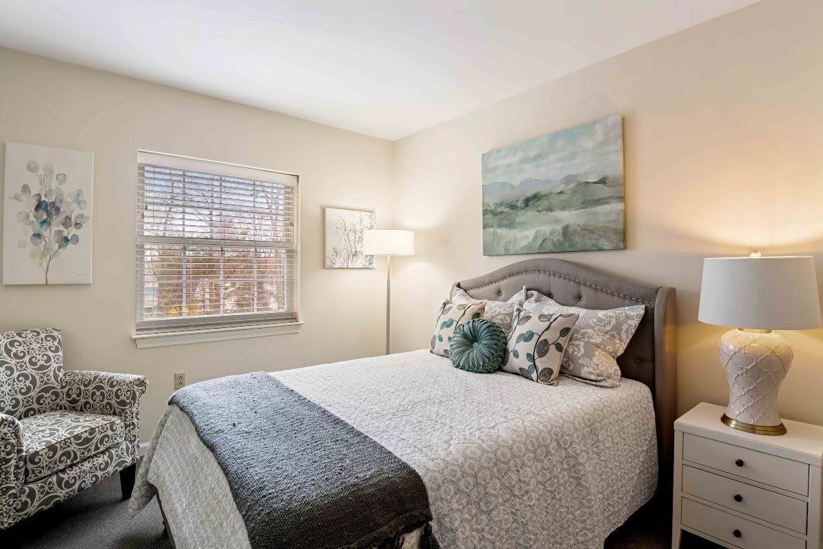 A cozy bedroom featuring a neatly made bed with decorative pillows, a standing lamp, and framed artwork on the walls.