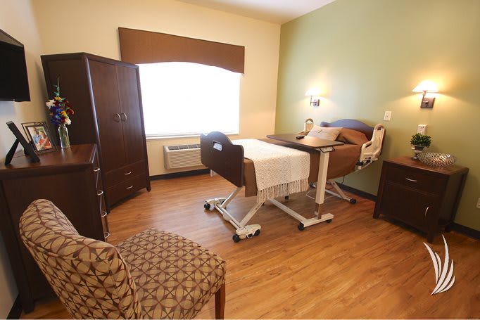 A cozy nursing home bedroom with a hospital bed, armchair, and wooden floors.