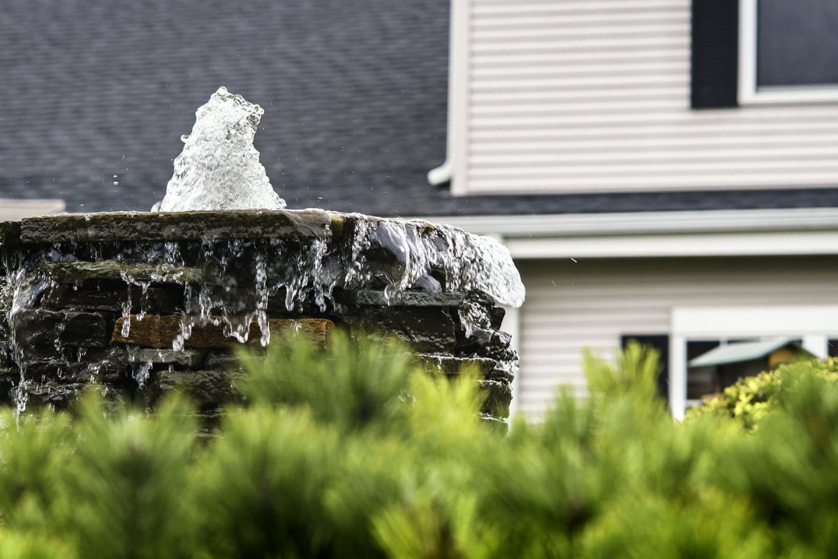 A bubbling stone fountain in front of a house surrounded by greenery.