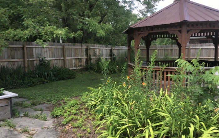 A tranquil garden featuring a wooden gazebo surrounded by lush greenery and enclosed by a fence.