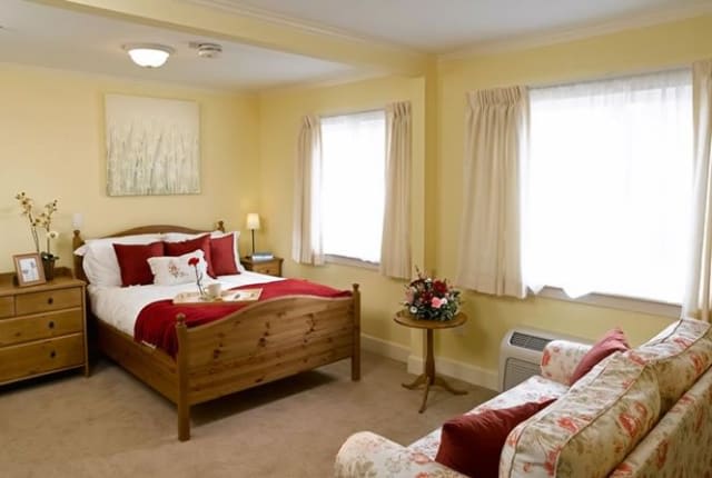 A cozy bedroom with a wooden bed, warm lighting, and soft yellow walls, featuring two windows with curtains.
