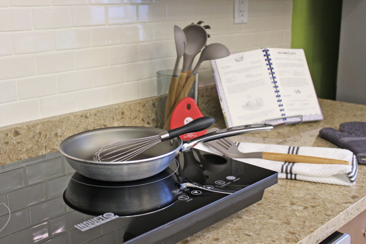 A kitchen countertop featuring an induction stove with a frying pan and whisk, accompanied by cooking utensils and an open cookbook.