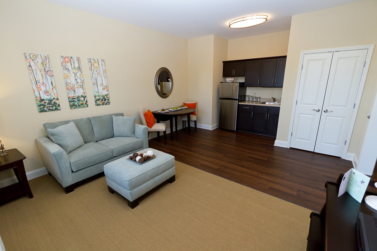 A cozy apartment living room with a sofa, ottoman, and a small dining area next to the kitchen.