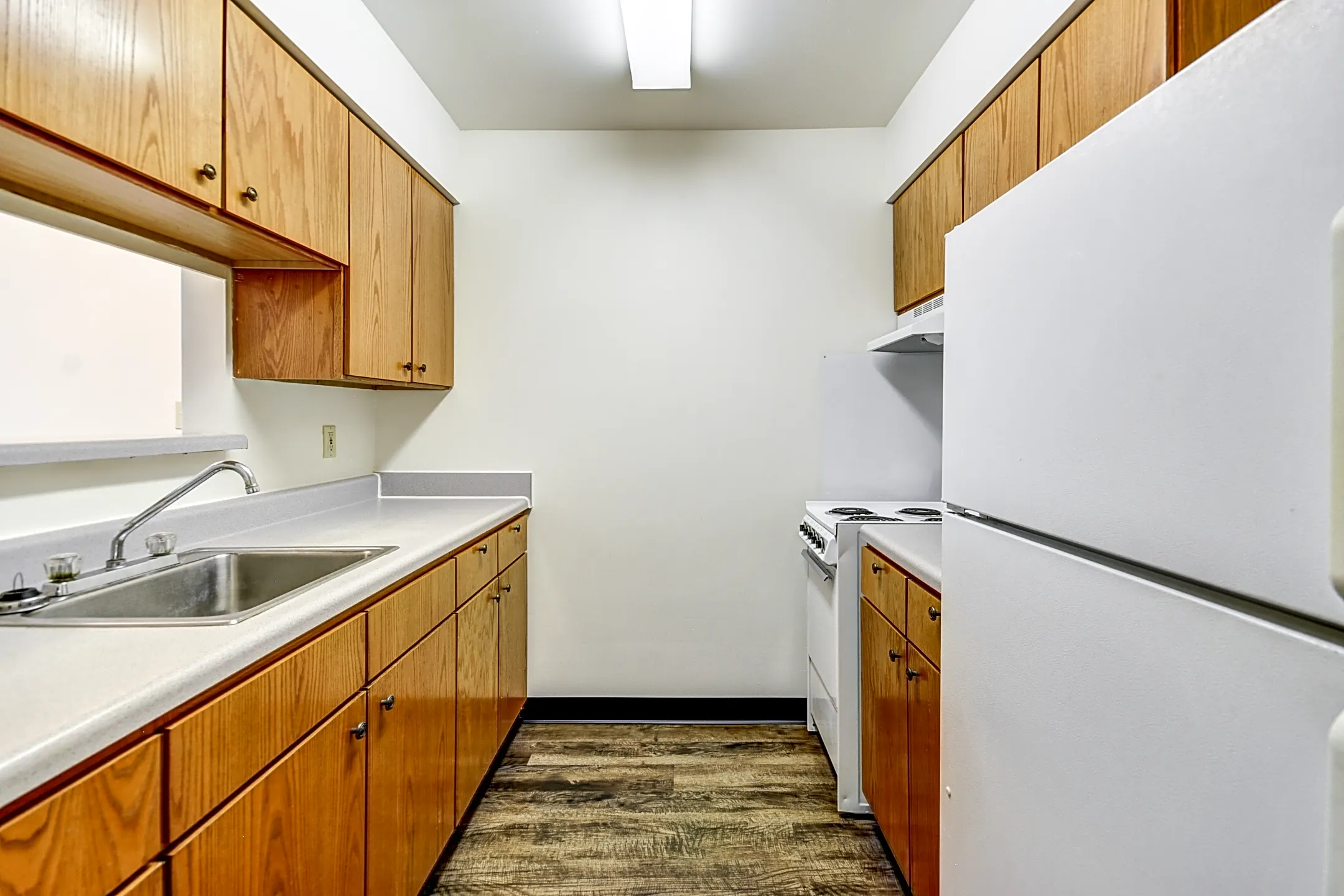 A narrow apartment kitchen with wood cabinets and white appliances.