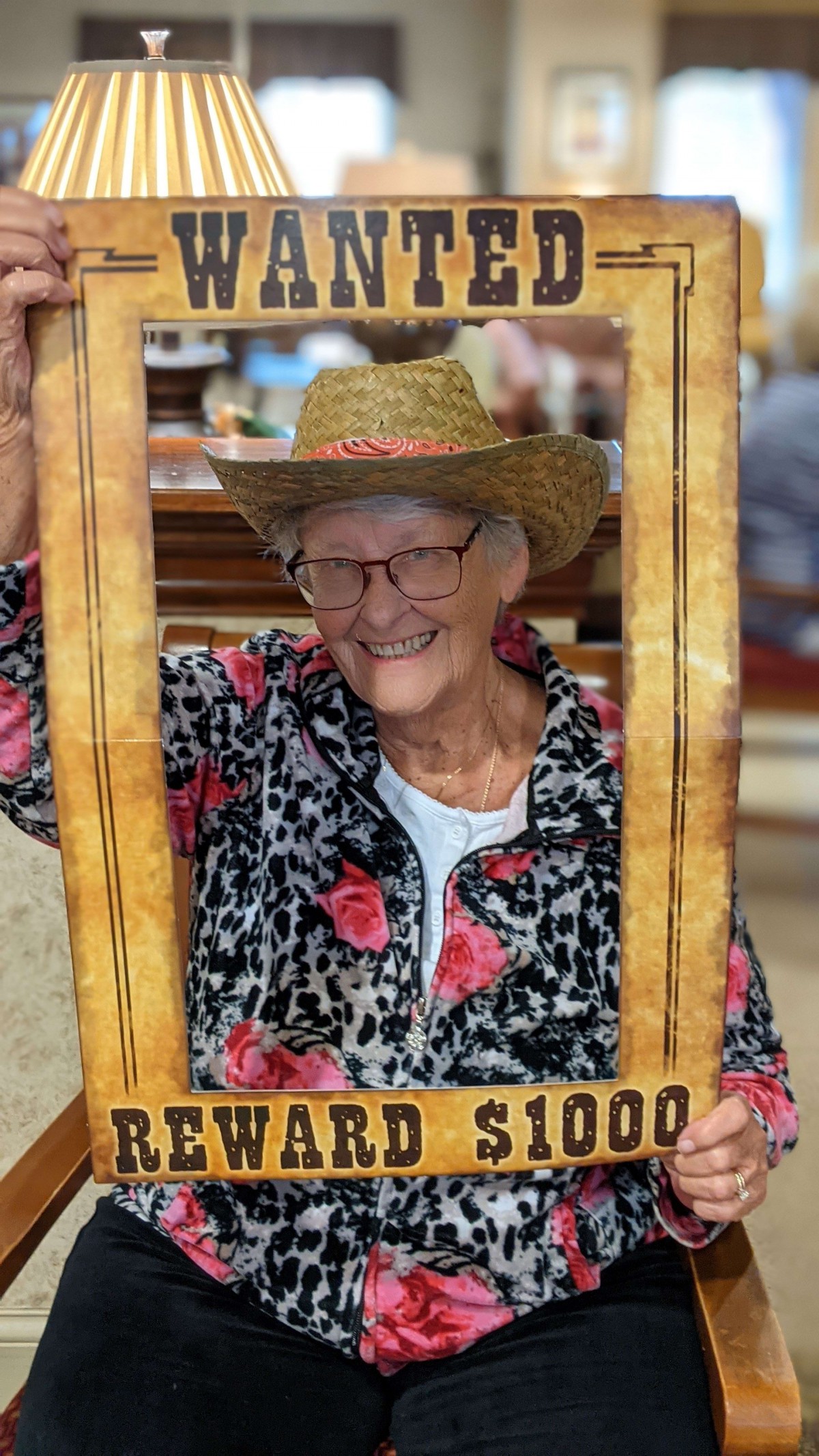 An elderly woman smiles while wearing a hat and posing with a wooden frame that resembles a reward poster.