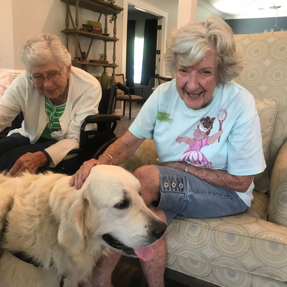 Two elderly women pet a dog while sitting indoors, smiling happily.