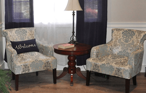 A cozy living room corner featuring two patterned chairs flanking a wooden side table with a lamp.