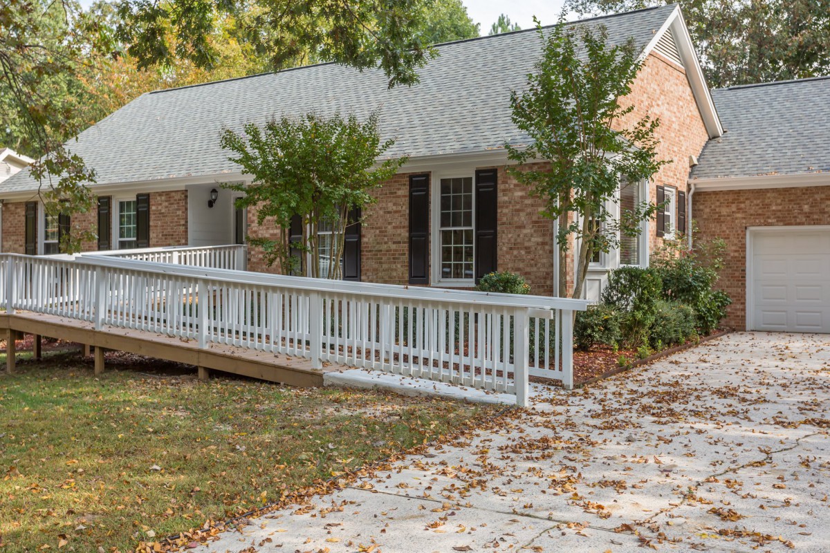 A single-story brick house with a white accessibility ramp surrounded by trees and fall leaves.