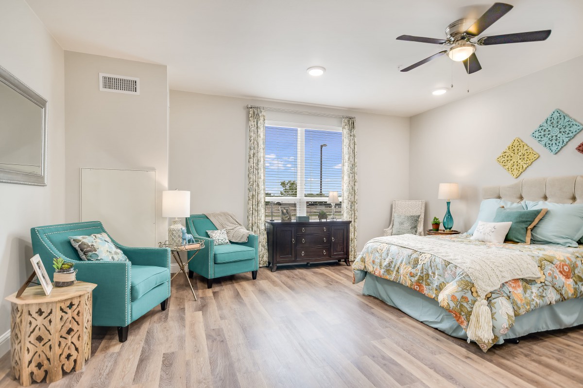 A modern and cozy bedroom featuring teal armchairs, a bed with floral bedding, and abundant natural light.