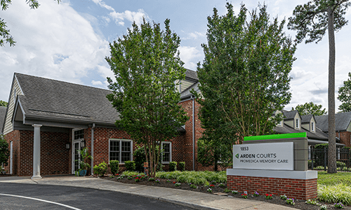 A brick office building surrounded by trees, featuring a sign at the entrance under a cloudy sky.