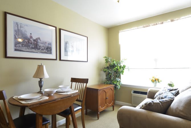 A cozy dining area with a table set, chairs, artwork on the wall, and natural light from a window.