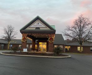 A brick building with a well-lit entrance surrounded by trees and a parking lot at dusk.